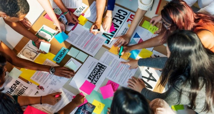 Funcionários debruçados em cima de uma mesa analisando planos estratégicos e criativos.