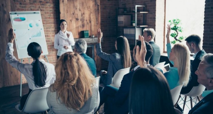 Executivos sentados numa sala de aula in company levantam à mão para questionar a treinadora.