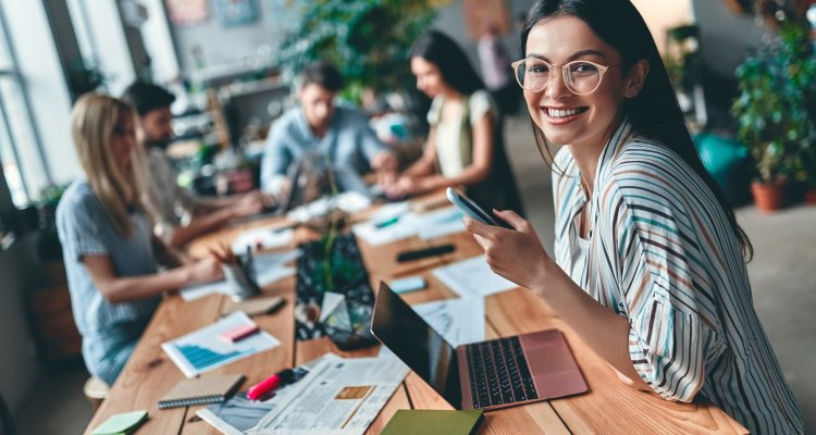 Executiva com óculos segura o celular na mão e a sua frente tem notebook, ao lado colegas de trabalho se reúnem.