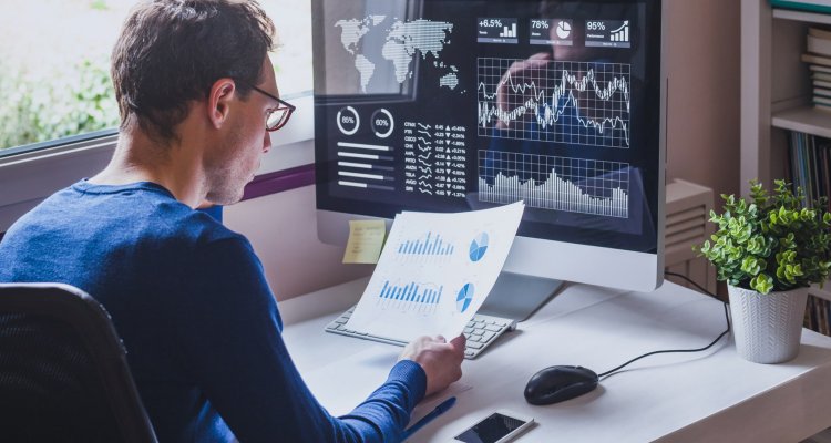 Um executivo sentado à sua mesa de trabalho observando um dashboard no papel e no monitor de seu computador